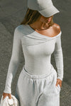 Front view of model posing outside in the street in the fitted pearl grey rib Off Shoulder Twist Long Sleeve top asymmetrical neckline with a twist detail and off-shoulder design