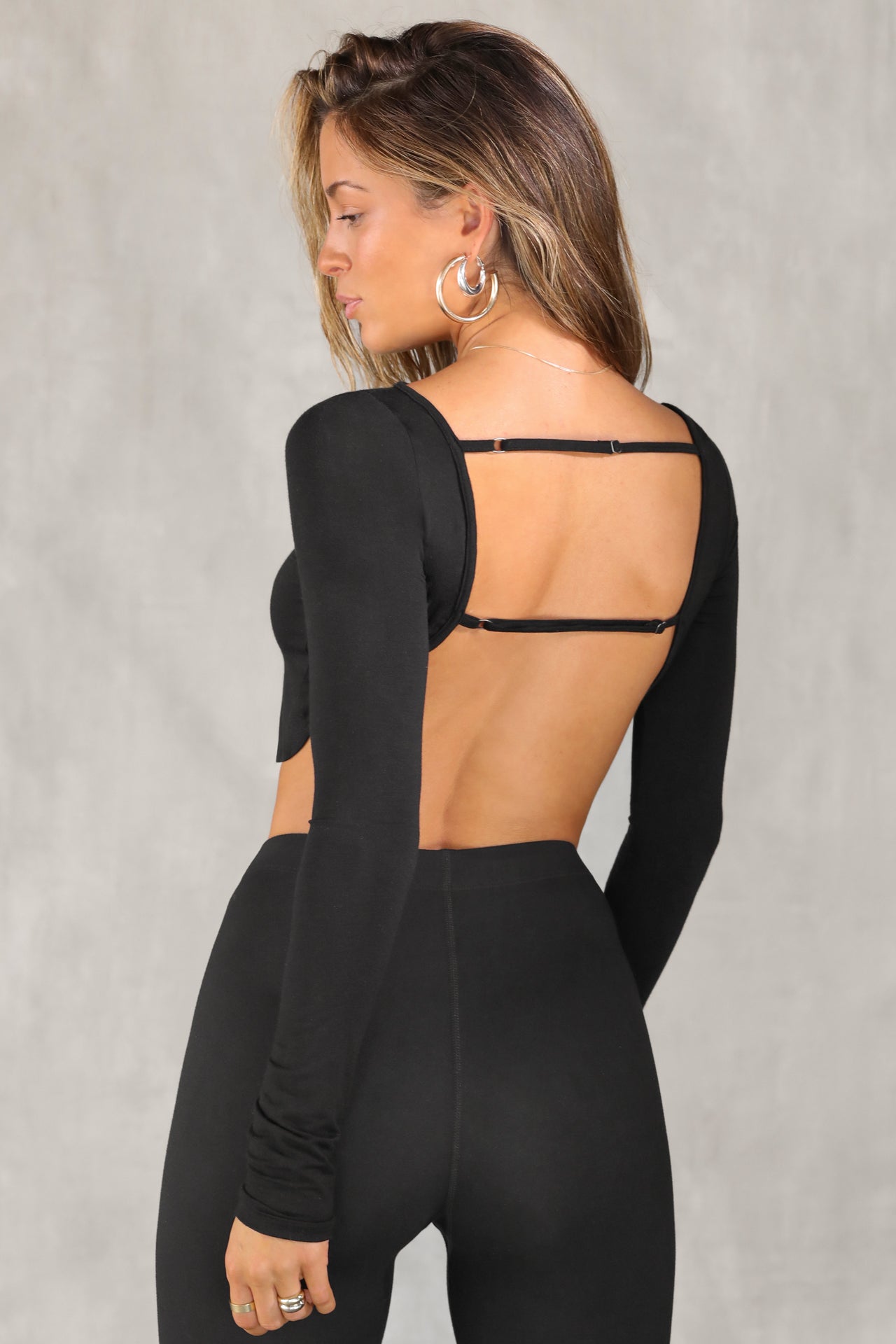 Back view of model posing in the fitted and cropped black modal Open Boatneck Long Sleeve top with a high neckline,curved hem, open back and adjustable strap details