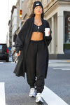 Full body front view of model posing outside in the street in the knee-length loose fitting black french terry Oversized Cardigan with a rolled neckline and extra long sleeves