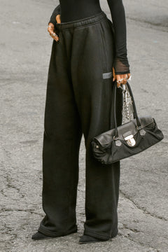 Side view of model from the waist down posing outside in the street in the relaxed fit black french terry Oversized Straight Leg Jogger with an elastic waistband, hidden drawstring, pocket details. and adjustable cinching at the ankle cuffs