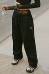 Front view of model from the waist down posing outside in the street in the relaxed fit black french terry Oversized Straight Leg Jogger with an elastic waistband, hidden drawstring, pocket details. and adjustable cinching at the ankle cuffs