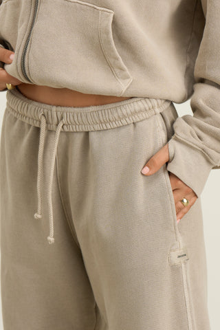 Close up detail view of model from the waist down posing in the relaxed fit sand french terry Oversized Straight Leg Jogger with an elastic waistband, hidden drawstring, pocket details. and adjustable cinching at the ankle cuffs