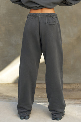 Back view of model from the waist down posing in the relaxed fit washed black french terry Oversized Straight Leg Jogger with an elastic waistband, hidden drawstring, pocket details. and adjustable cinching at the ankle cuffs