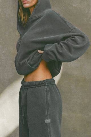 Side view of model posing in the relaxed fit washed black french terry Oversized Straight Leg Jogger with an elastic waistband, hidden drawstring, pocket details,  adjustable cinching at the ankle cuffs and a custom Joah Brown logo patch below the pocket