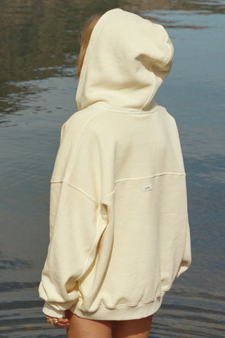 Back view of model posing outside by a lake in the relaxed fit buttercream french terry Classic Zip Hoodie with a full length front zip closure, side pockets and thumbholes in the fitted cuffs