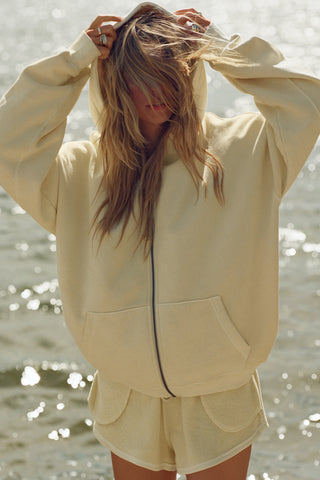 Front view of model posing outside by a lake in the relaxed fit buttercream french terry Classic Zip Hoodie with a full length front zip closure, side pockets and thumbholes in the fitted cuffs