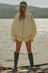 Full body front view of model posing outside by a lake in the relaxed fit buttercream french terry Classic Zip Hoodie with a full length front zip closure, side pockets and thumbholes in the fitted cuffs