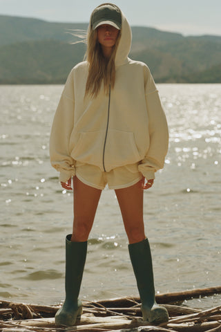 Full body front view of model posing outside by a lake in the relaxed fit buttercream french terry Classic Zip Hoodie with a full length front zip closure, side pockets and thumbholes in the fitted cuffs
