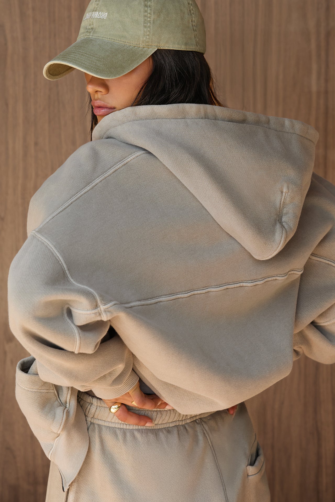 Back view of model posing in the oversized comfortable sand french terry Empire Zip Hoodie with a full length front zip closure, side pockets and thumbholes in the fitted cuffs