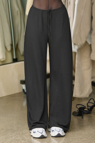 Front view of model from the waist down posing in the relaxed fit black modal Relaxed Straight Leg Pant with a wide leg, elastic waistband, adjustable drawstring tie and subtle pleating at the sides.
