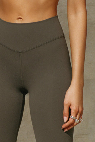 Close up detail front view of model from the waist down posing in the full length bootcut sueded carob Second Skin Bootcut Legging with a wide v-shaped waistband and split detail at the leg opening