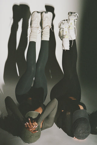 Two models lying on their back with their legs up in the air wearing the full length and high-waisted sueded essex and sueded onyx Second Skin Leggings with a wide, v-shaped waistband