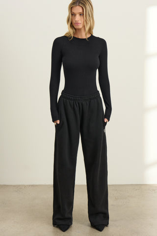 Full body front view of model posing in fitted black flexrib Sheer Back Long Sleeve top with a crew neckline and sheer mesh back panel.