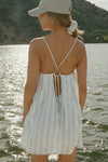 Back view of model posing outside by a lake in the lightweight natural white and blue stripe gauze Shift Mini Dress with a scoop neckline, front keyhole detail and open back with adjustable straps and a tie closure