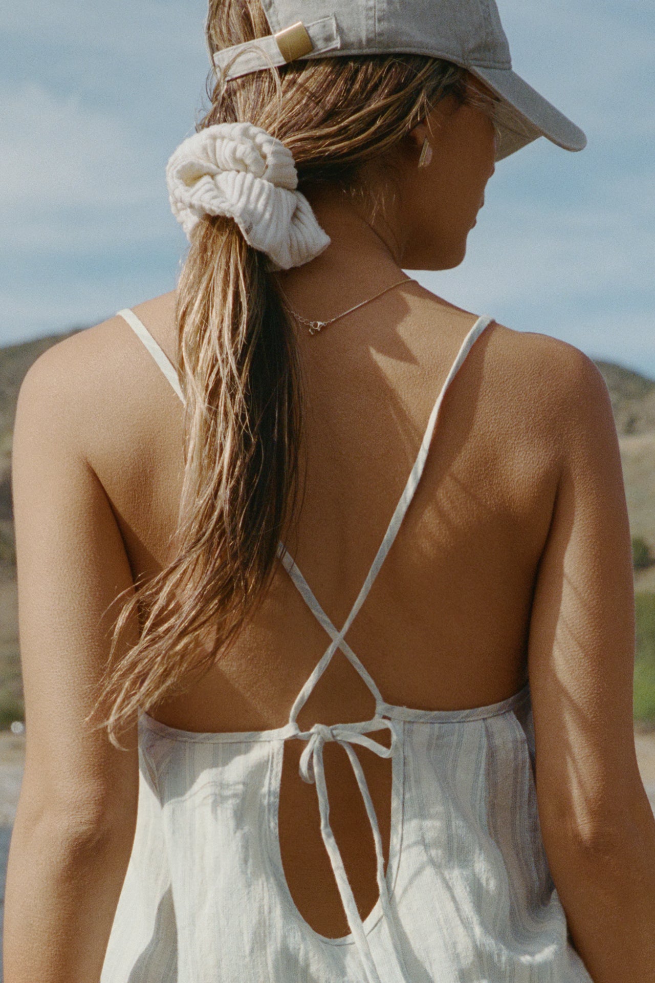 Close up back view of model posing outside by a lake in the lightweight natural white and blue stripe gauze Shift Mini Dress with a scoop neckline, front keyhole detail and open back with adjustable straps and a tie closure