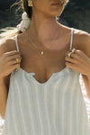 Close up front view of model posing outside by a lake in the lightweight natural white and blue stripe gauze Shift Mini Dress with a scoop neckline, front keyhole detail and open back with adjustable straps and a tie closure