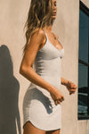 Side view of model posing in the form fitting stretchy pearl grey rib mini Slip Dress with thin straps, a u neckline and a tiny side slit at the hem