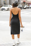 Full body back view of model posing in the stretchy form fitting black rib Strappy Slit Dress with a high front slit, thin corded straps and a low scoop back that hits at mid-calf