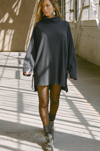 Full body front view of model posing in the relaxed and slouchy tunic-style midnight luxe knit Turtleneck Tunic with dropped shoulders, side slits and adjustable ties for a customized shape