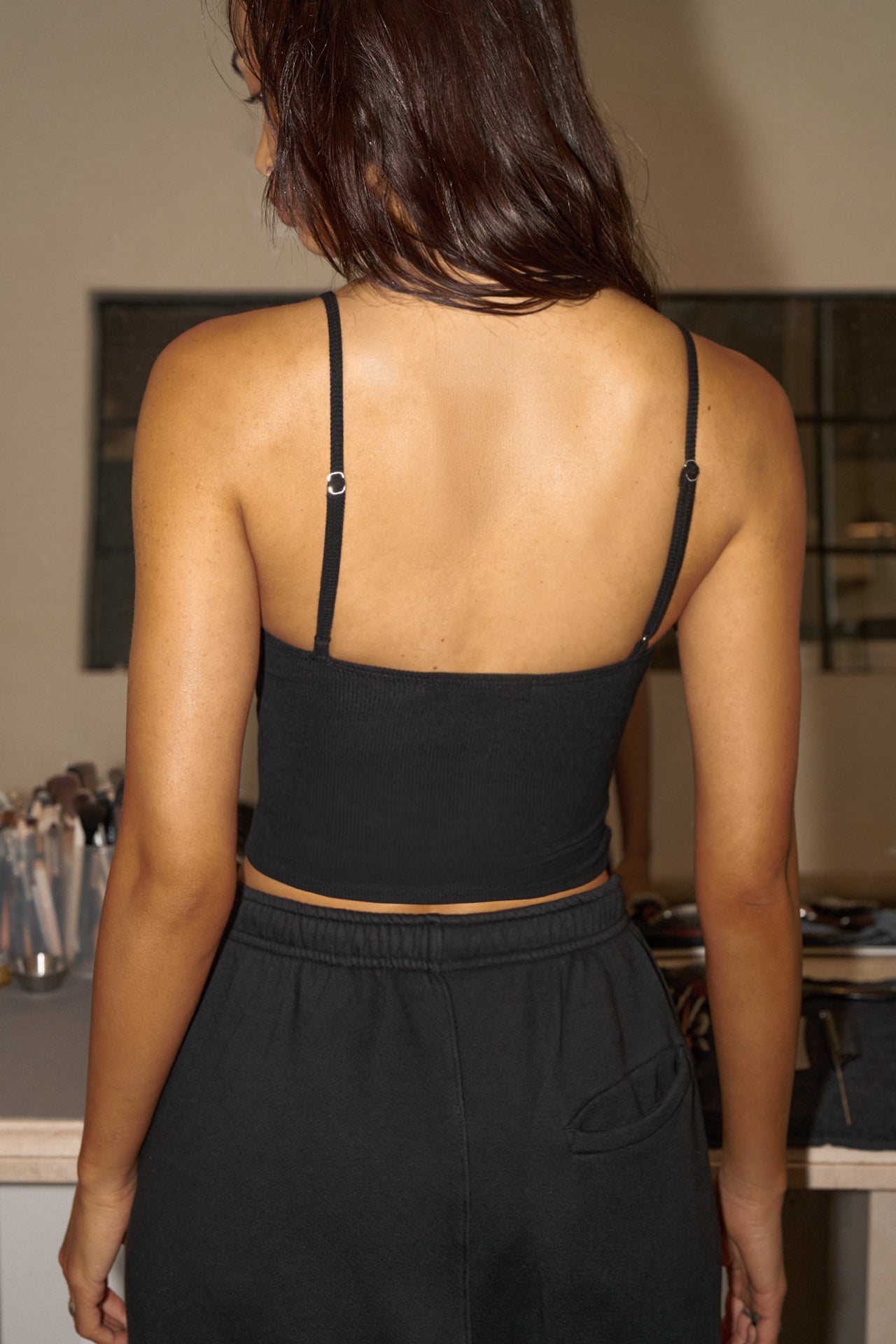 Back view of model posing in the fitted and slightly cropped black flexrib Strappy Scoop Tank with a scoop neckline and adjustable straps. 