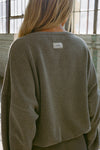 Back view of model posing in the slouchy oakmoss sherpa V Neck Pullover sweatshirt with a wide V-neckline that can be worn on or off the shoulders and a custom Joah Brown logo patch on the back