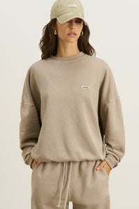 Front view of model posing in the relaxed fit sand french terry Varsity Crew Pullover sweatshirt with a crew neckline, dropped shoulders and a Joah Brown logo patch at the chest