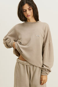 Front view of model posing in the relaxed fit sand french terry Varsity Crew Pullover sweatshirt with a crew neckline, dropped shoulders and a Joah Brown logo patch at the chest