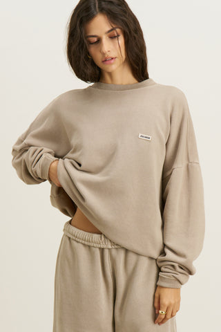 Front view of model posing in the relaxed fit sand french terry Varsity Crew Pullover sweatshirt with a crew neckline, dropped shoulders and a Joah Brown logo patch at the chest