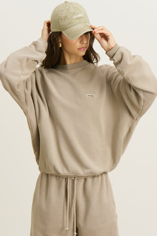 Front view of model posing in the relaxed fit sand french terry Varsity Crew Pullover sweatshirt with a crew neckline, dropped shoulders and a Joah Brown logo patch at the chest