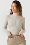 Front view of model posing in the fitted dune flexrib Stitch Mock Neck Long Sleeve top with tonal stitching on the front, a mock neckline and thumbholes in the cuffs