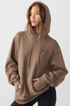 Front view of model posing in the oversized comfortable cocoa french terry with logo Empire Pullover Hoodie sweatshirt with a Joah Brown logo patch at the front left chest, kangaroo pocket and drawstrings at the bottom of the hood