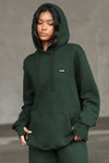 Front view of model posing in the oversized comfortable pine french terry with logo Empire Pullover Hoodie sweatshirt with a Joah Brown logo patch at the front left chest, kangaroo pocket and drawstrings at the bottom of the hood