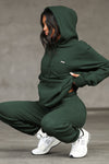 Full body side view of model posing in a crouching position wearing the oversized comfortable pine french terry with logo Empire Pullover Hoodie sweatshirt with a Joah Brown logo patch at the front left chest, kangaroo pocket and drawstrings at the bottom of the hood