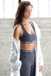 Side view of model posing in front of a window wearing the fitted and stretchy sueded navy The Sports Bra with a scoop neckline and an elastic under band printed with the Joah Brown logo