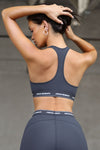 Back view of model posing the fitted and stretchy sueded navy The Sports Bra with a racerback and an elastic under band printed with the Joah Brown logo