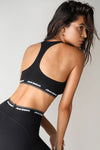 Back view of model posing the fitted and stretchy sueded onyx The Sports Bra with a racerback and an elastic under band printed with the Joah Brown logo