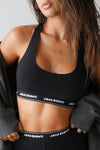 Close up front view of model posing the fitted and stretchy sueded onyx The Sports Bra with a scoop neckline and an elastic under band printed with the Joah Brown logo