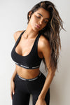Front view of model posing the fitted and stretchy sueded onyx The Sports Bra with a scoop neckline and an elastic under band printed with the Joah Brown logo